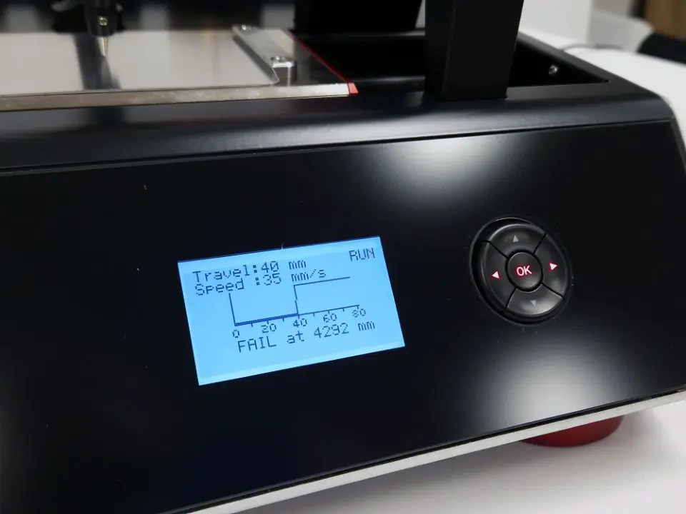 Black printer front panel with a blue backlit LCD showing travel and speed readouts and a small graph.