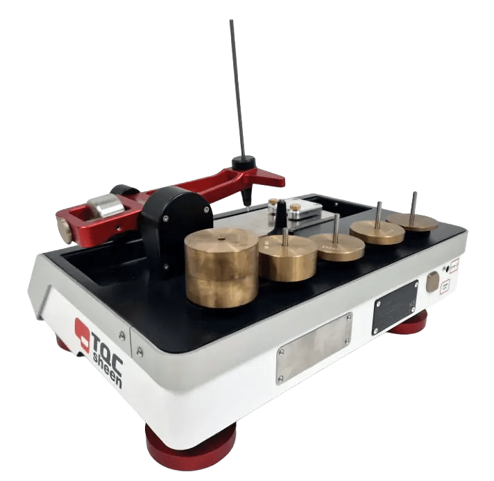 Laboratory force-testing device with brass weights on a black panel and a red lever arm attached to a white base with a dark top surface.