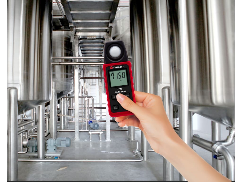 Hand holding a red light meter in a stainless-steel industrial facility, the display reads 71.50 Fc.